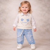 Child wearing a white sweater with blue floral designs and light blue jeans on a neutral background